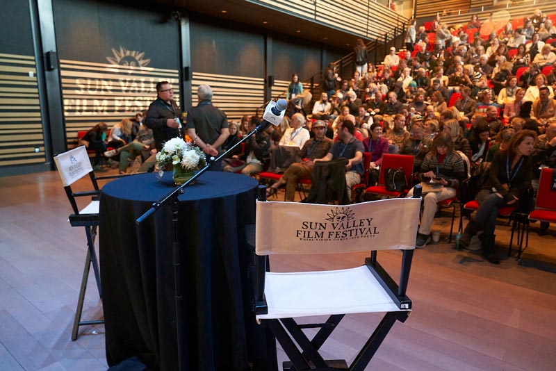 A directors chair besides a table on stage with a crowd in the background